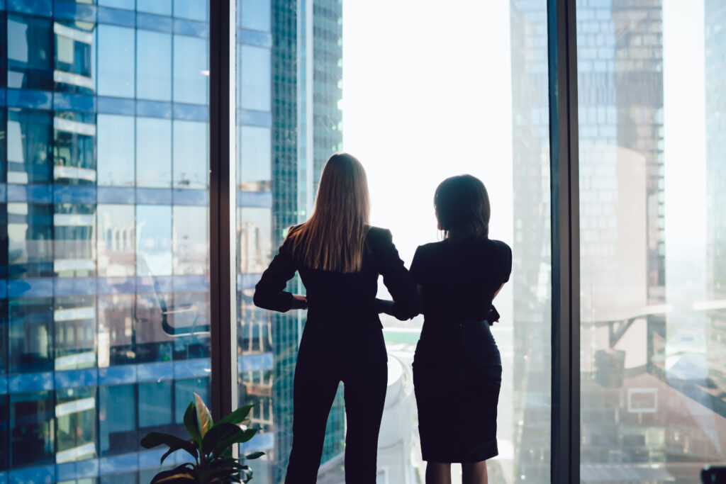 back view of female colleagues in formal wear standing near window looking at modern exterior of skyscrapers in business center, prosperous women standing together planning prosperous future success