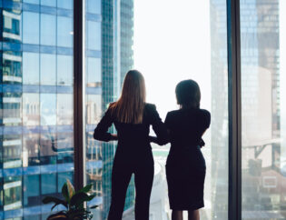 back view of female colleagues in formal wear standing near window looking at modern exterior of skyscrapers in business center, prosperous women standing together planning prosperous future success