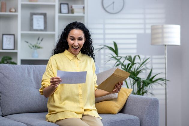 joyful and happy woman at home received mail envelope with notification, hispanic woman reading good news on paper and happy sitting on sofa in living room